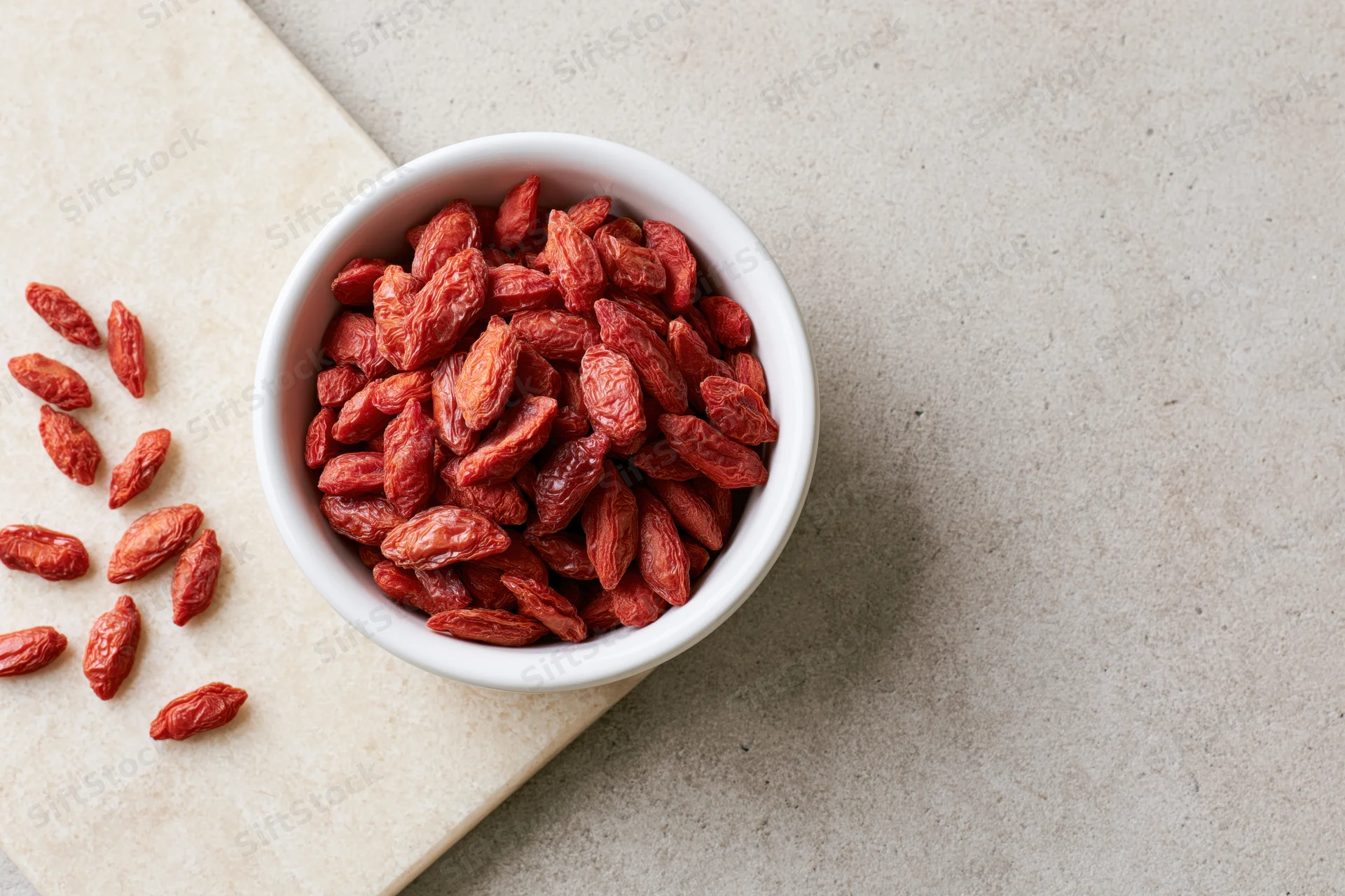 Dried goji berries in a white ceramic bowl on a stone countertop with scattered berries beside it