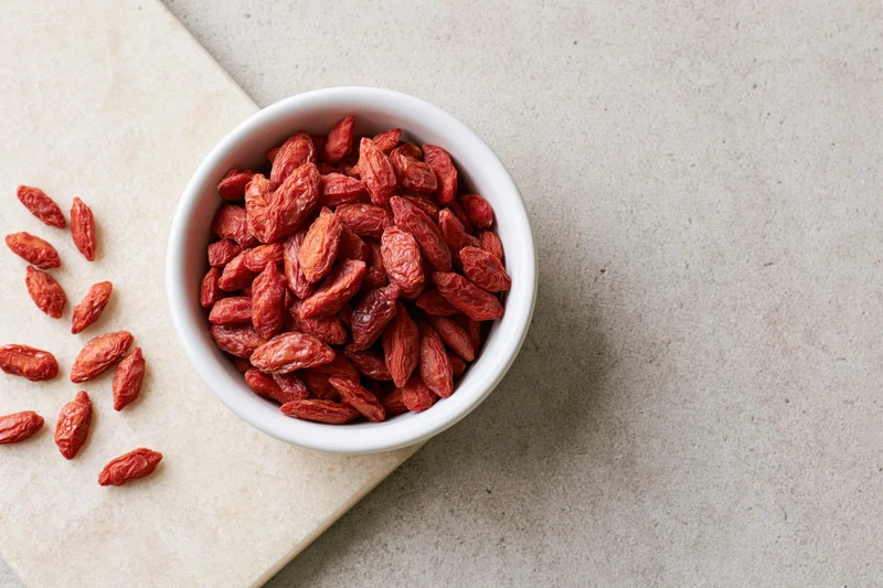 Dried goji berries in a white ceramic bowl on a stone countertop with scattered berries beside it