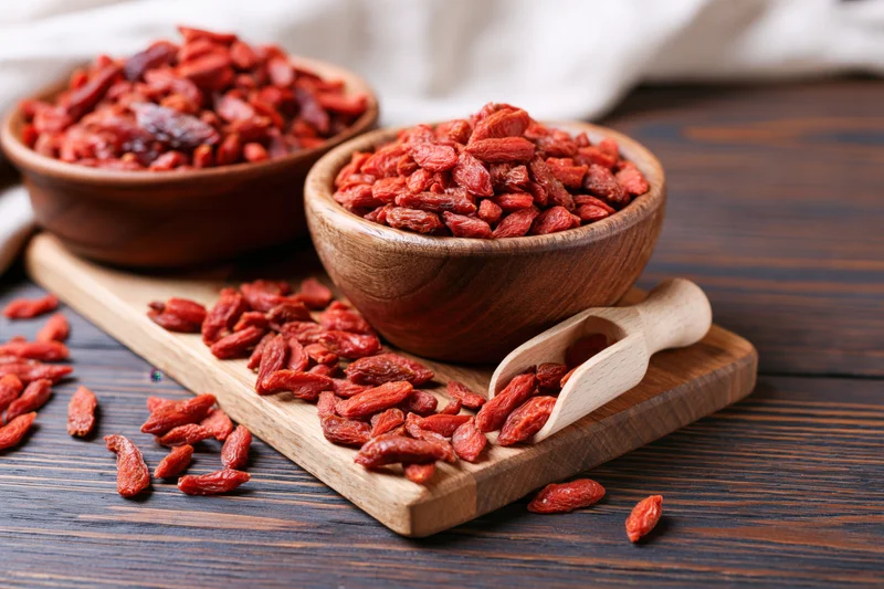 Dried goji berries in rustic wooden bowls on a dark wood table with a small wooden scoop and scattered berries around