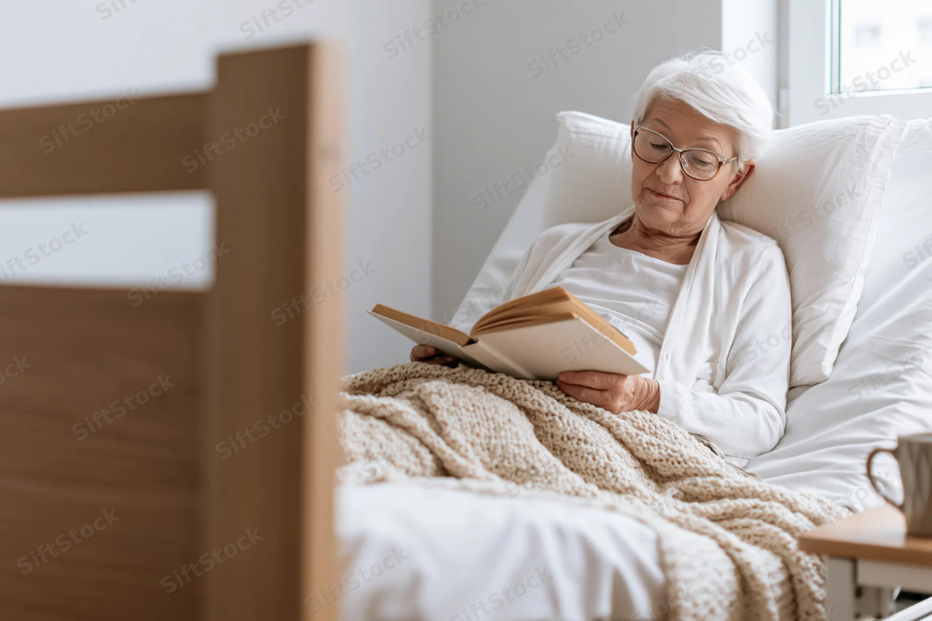 Elderly woman reading a book in bed with white bedding and a knit blanket