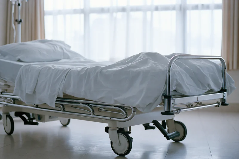 Empty adjustable hospital bed with white linens and chrome frame in a clinical room near a window