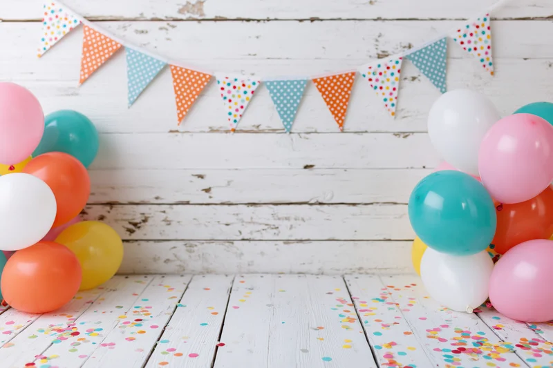 Colorful balloons and confetti arranged on white wooden floor with banner above, copyspace in center