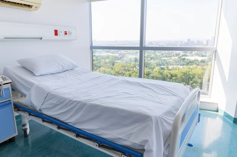 Empty hospital bed with white linens and a single pillow positioned near a large window overlooking a city and greenery