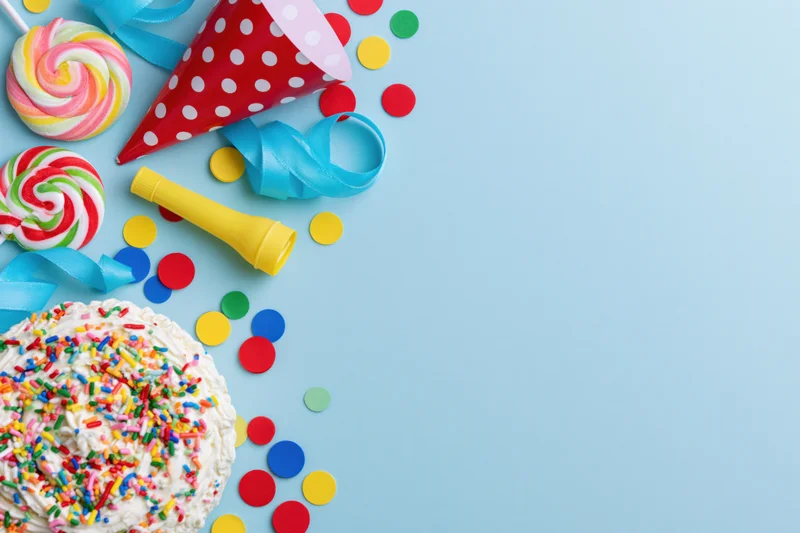 Birthday cake with sprinkles, lollipops, party hat and confetti arranged along left side on blue background