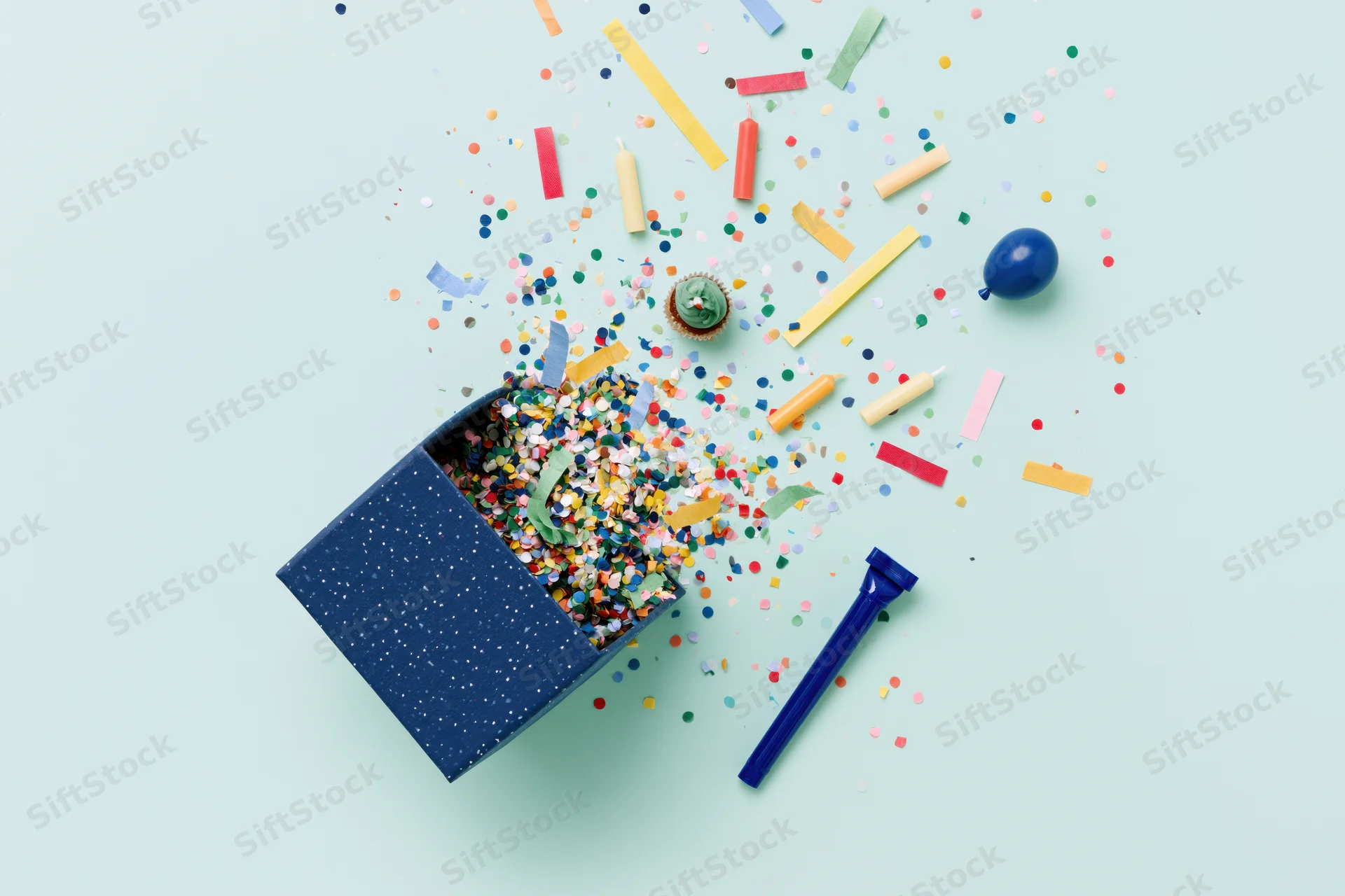 Birthday party items exploding from a box on a pastel background