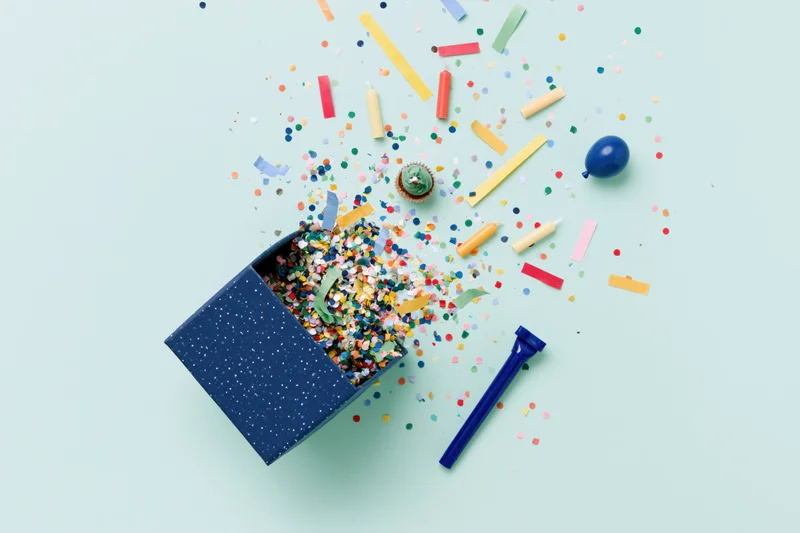 Birthday party items exploding from a box on a pastel background