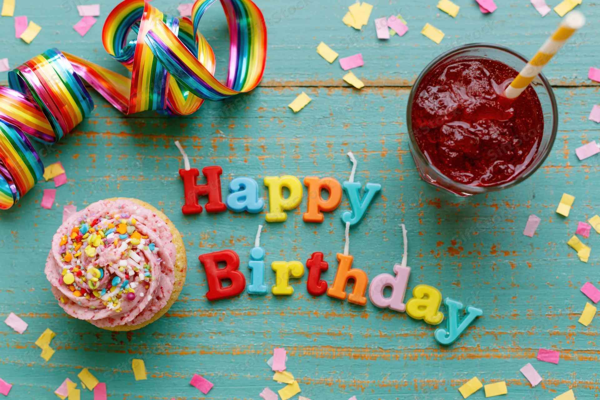 Colorful birthday party items arranged on a turquoise wooden table with cupcake, candles, streamers and confetti