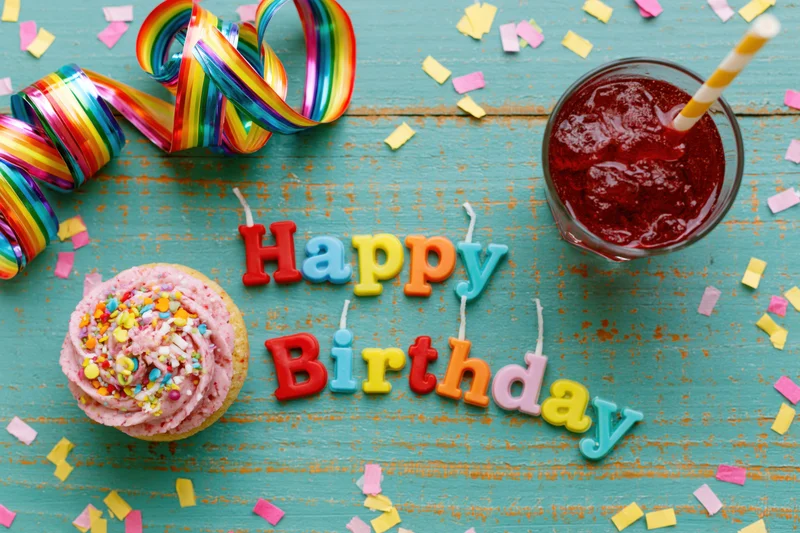 Colorful birthday party items arranged on a turquoise wooden table with cupcake, candles, streamers and confetti