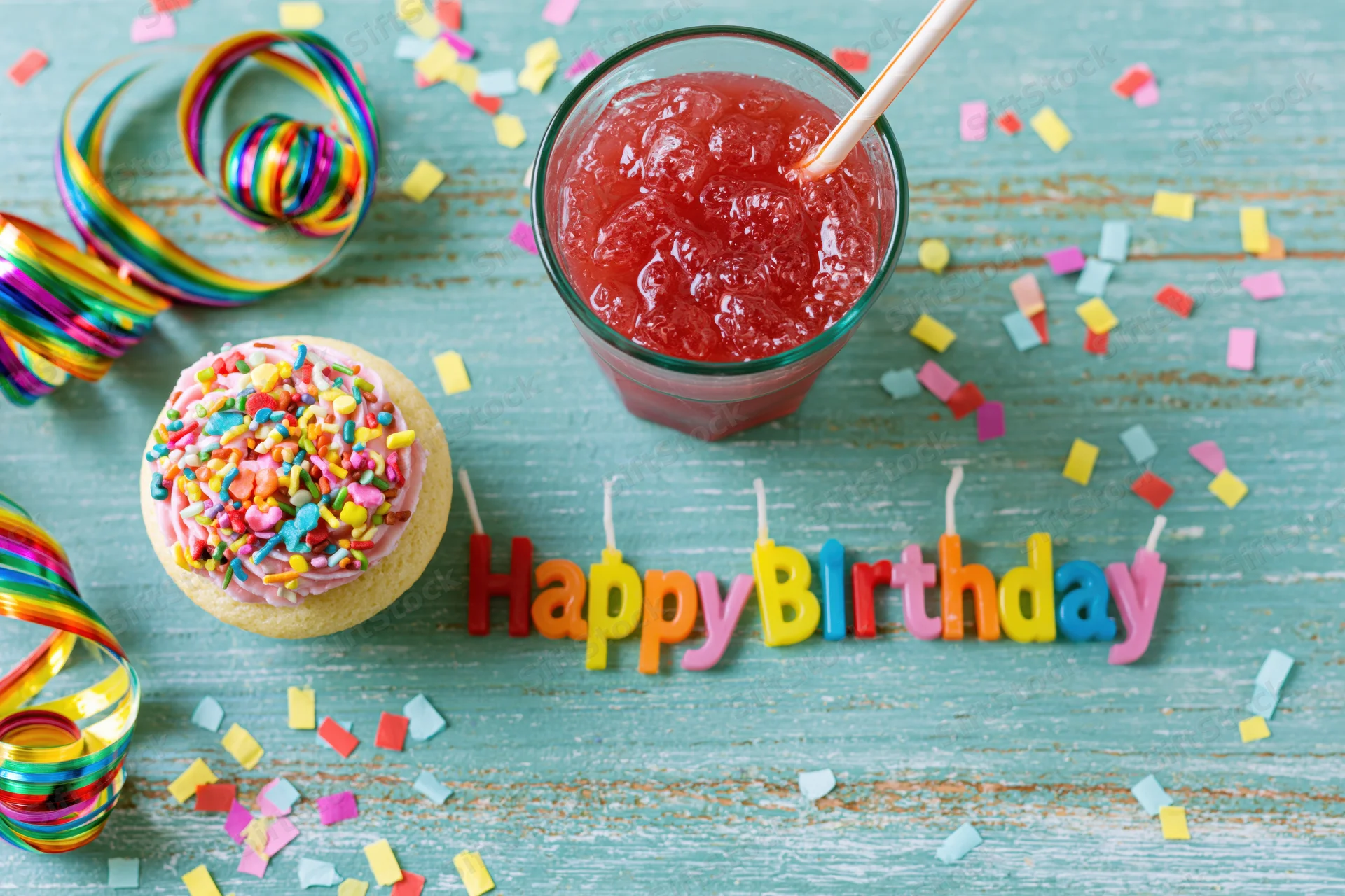 Colorful birthday party items arranged on a turquoise wooden surface with a cupcake, slushie, candles and confetti