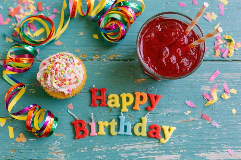 Colorful birthday party items arranged on a turquoise wooden surface with a cupcake, drink, and candy letters