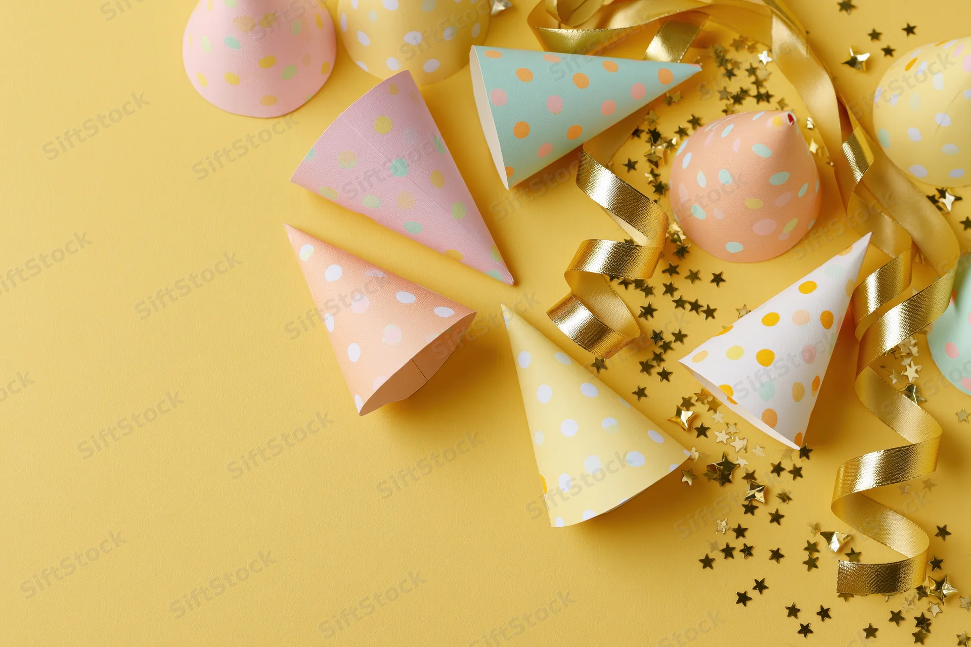 Colorful paper party hats and gold ribbon streamers arranged on yellow surface with confetti and copyspace