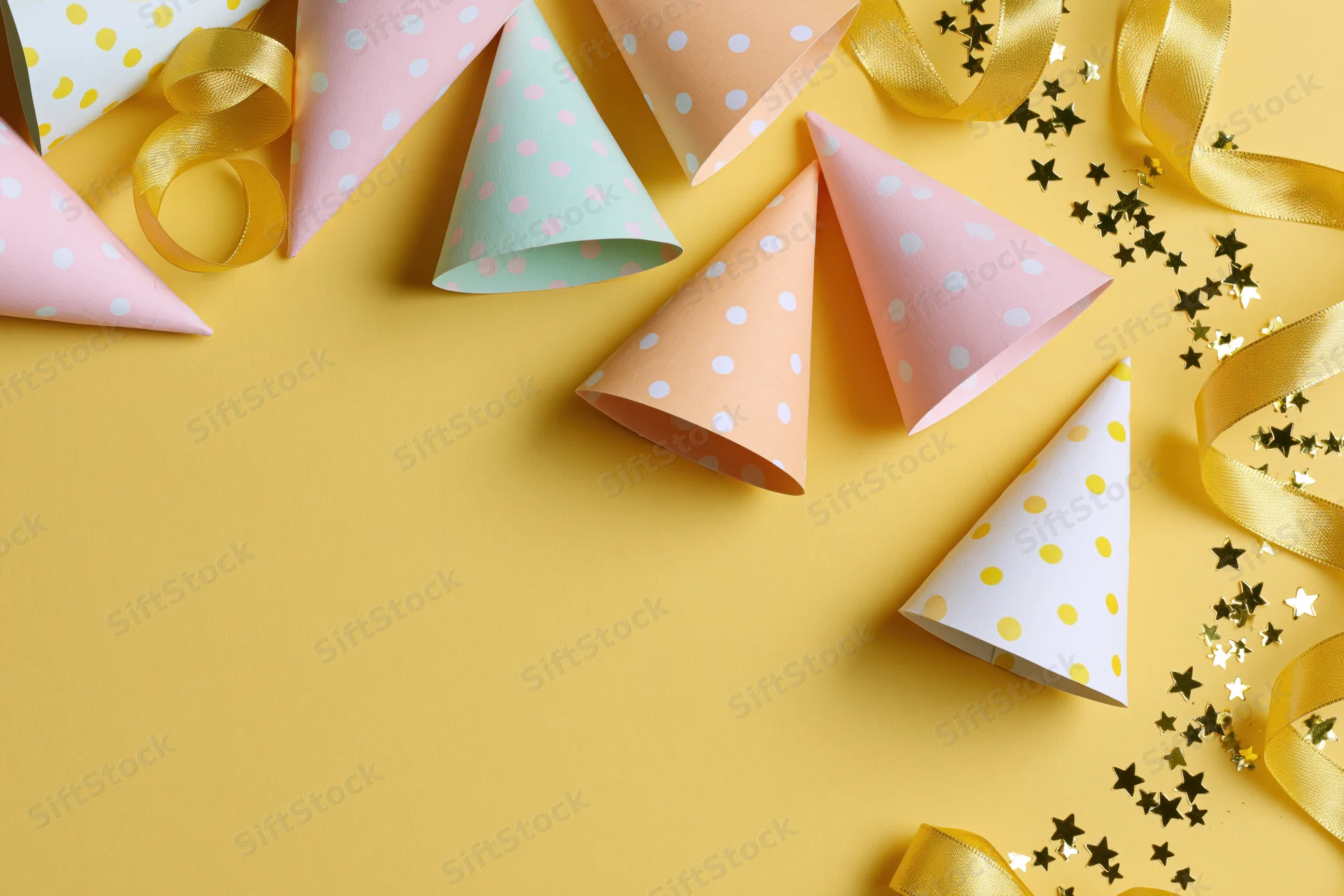 Colorful paper party hats and gold ribbon streamers arranged on yellow surface with star confetti