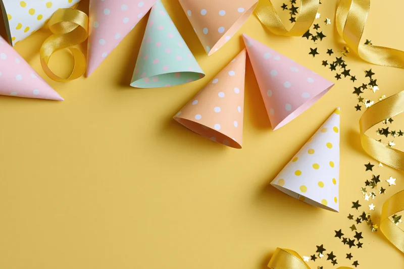 Colorful paper party hats and gold ribbon streamers arranged on yellow surface with star confetti