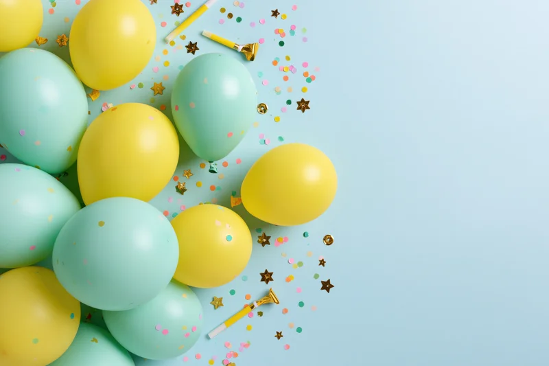 Mint green and yellow balloons with confetti and noisemakers arranged in flat lay on light blue surface
