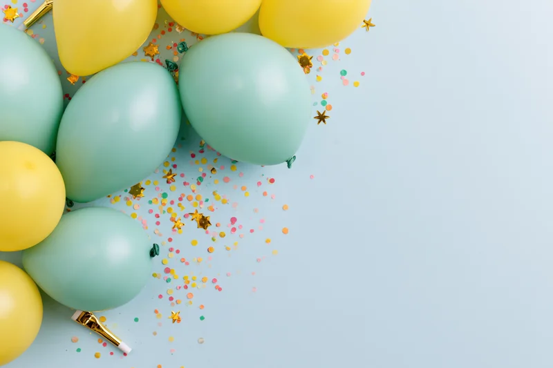 Mint green and yellow balloons with scattered confetti and gold stars on a light blue surface in a flat lay arrangement