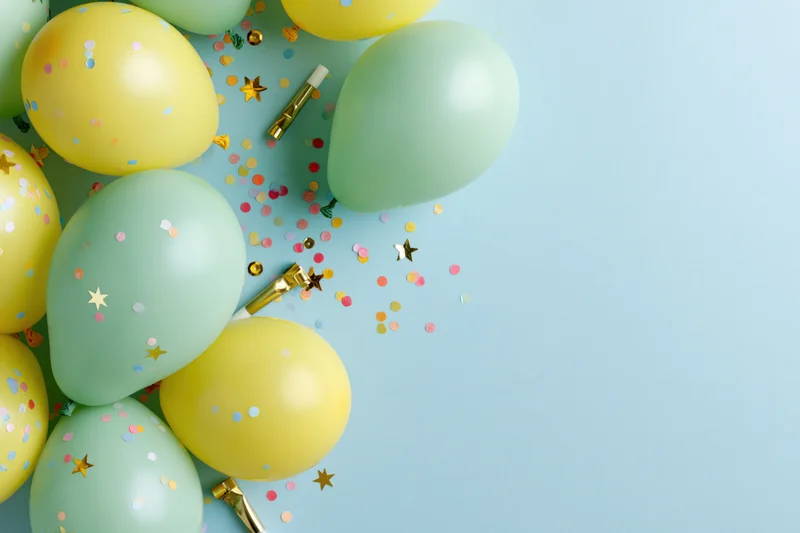 Mint green and yellow balloons with confetti and party blowers arranged in a flat lay on a light blue surface