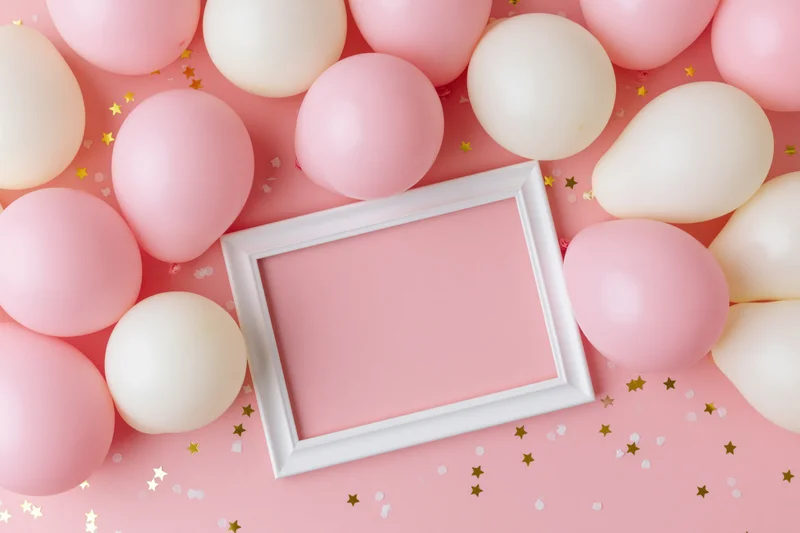 White frame mockup surrounded by pink and white balloons on a pink surface with gold star confetti, flat lay top view