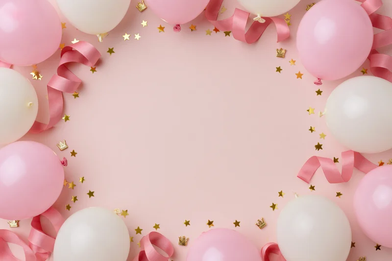 Pink and white balloons with ribbon streamers and gold confetti arranged in a circular frame on a pastel pink surface
