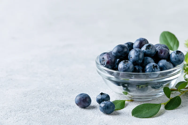 Fresh blueberries in a clear glass bowl on a white surface with scattered berries and green leaves