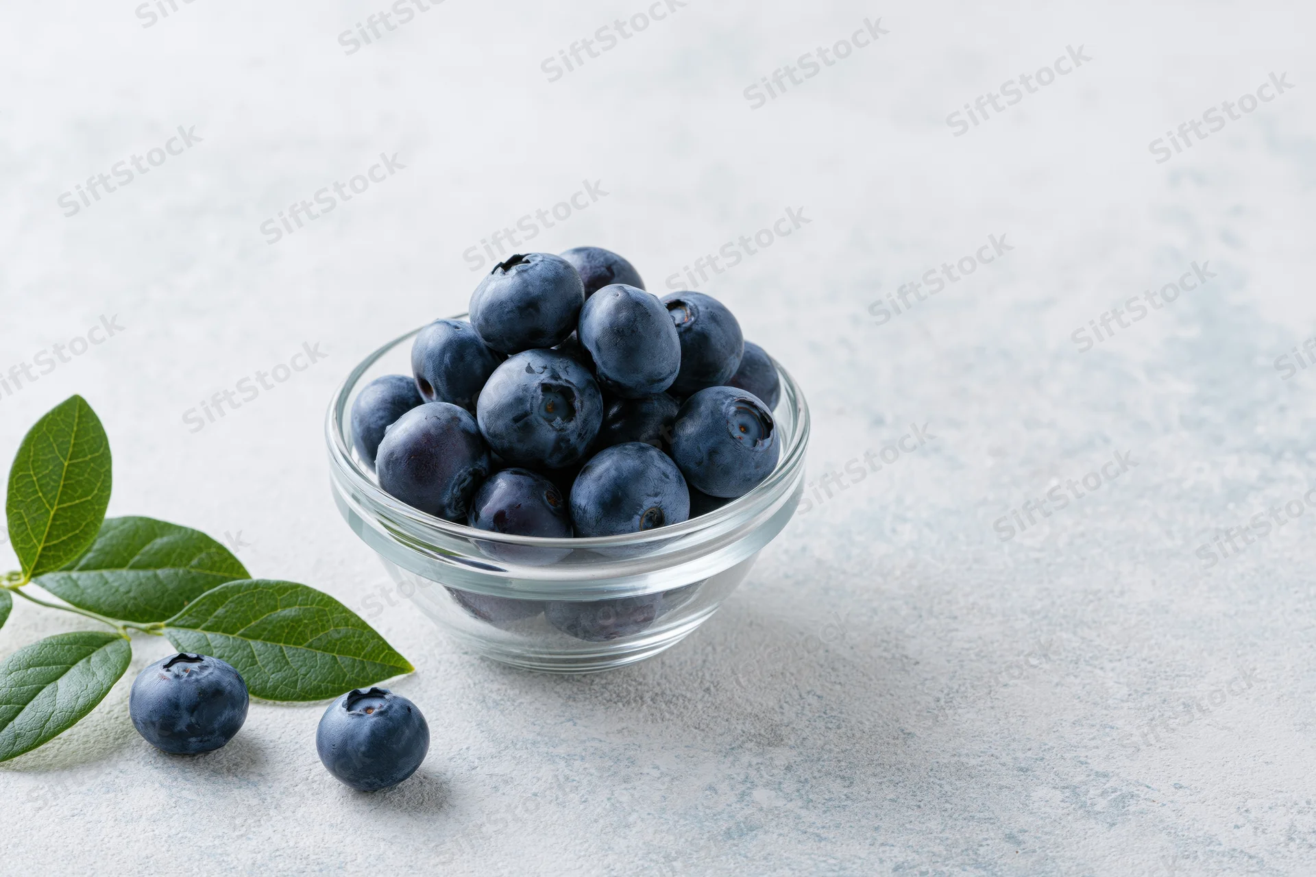 Fresh blueberries in a small glass bowl on a light surface with leaves and scattered berries nearby