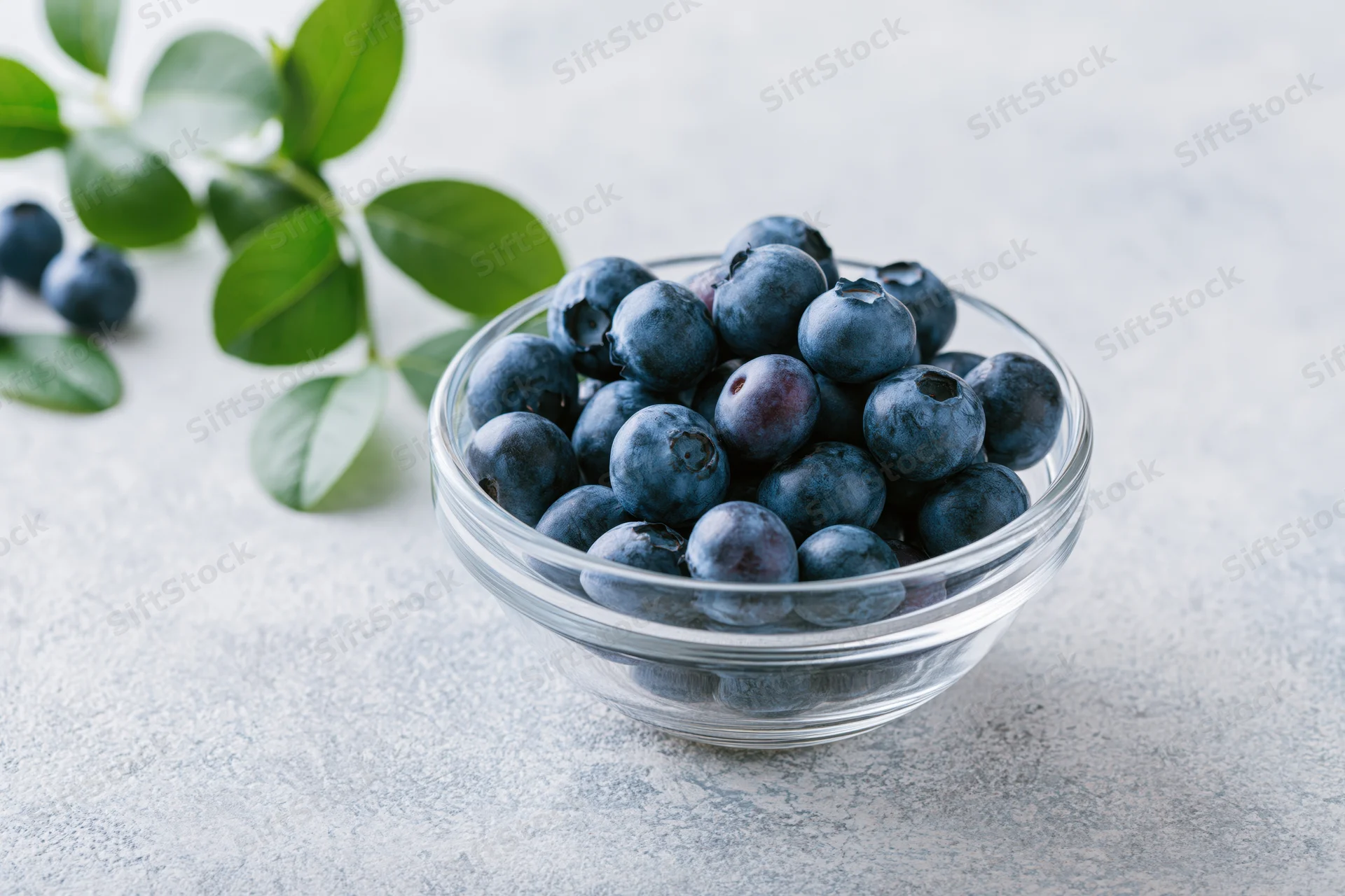 Fresh blueberries in a clear glass bowl on a light surface with green leaves nearby