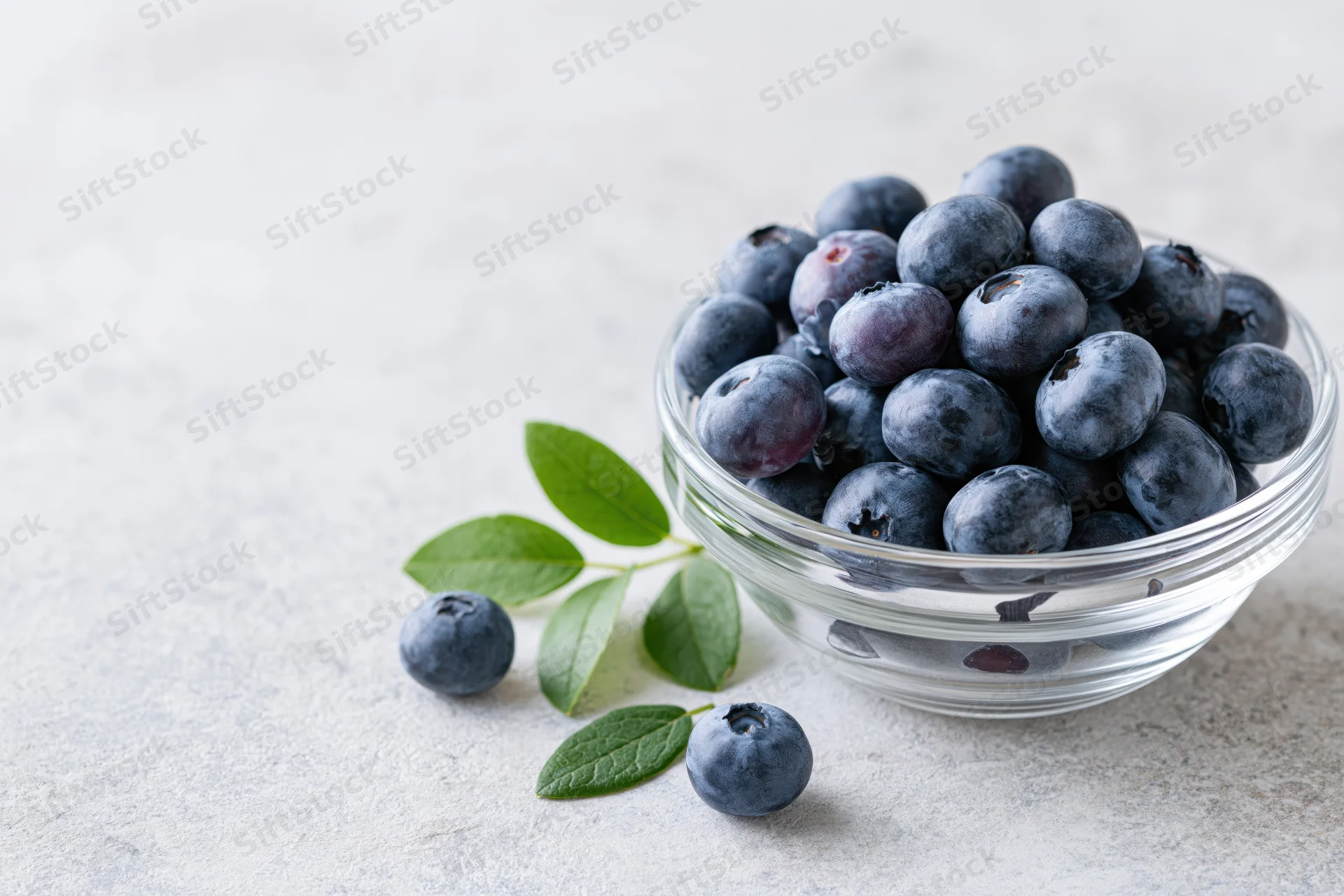 Fresh blueberries in a clear glass bowl on a light surface with leaves and scattered berries nearby