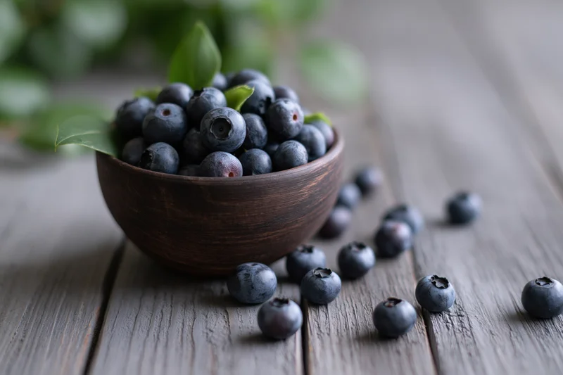 Fresh blueberries in a dark wooden bowl on a rustic wooden table with scattered berries and green leaves