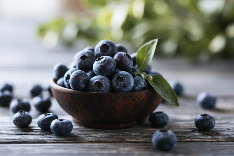 Fresh blueberries piled in a dark wooden bowl on a rustic wooden table with scattered berries and green leaves in the background