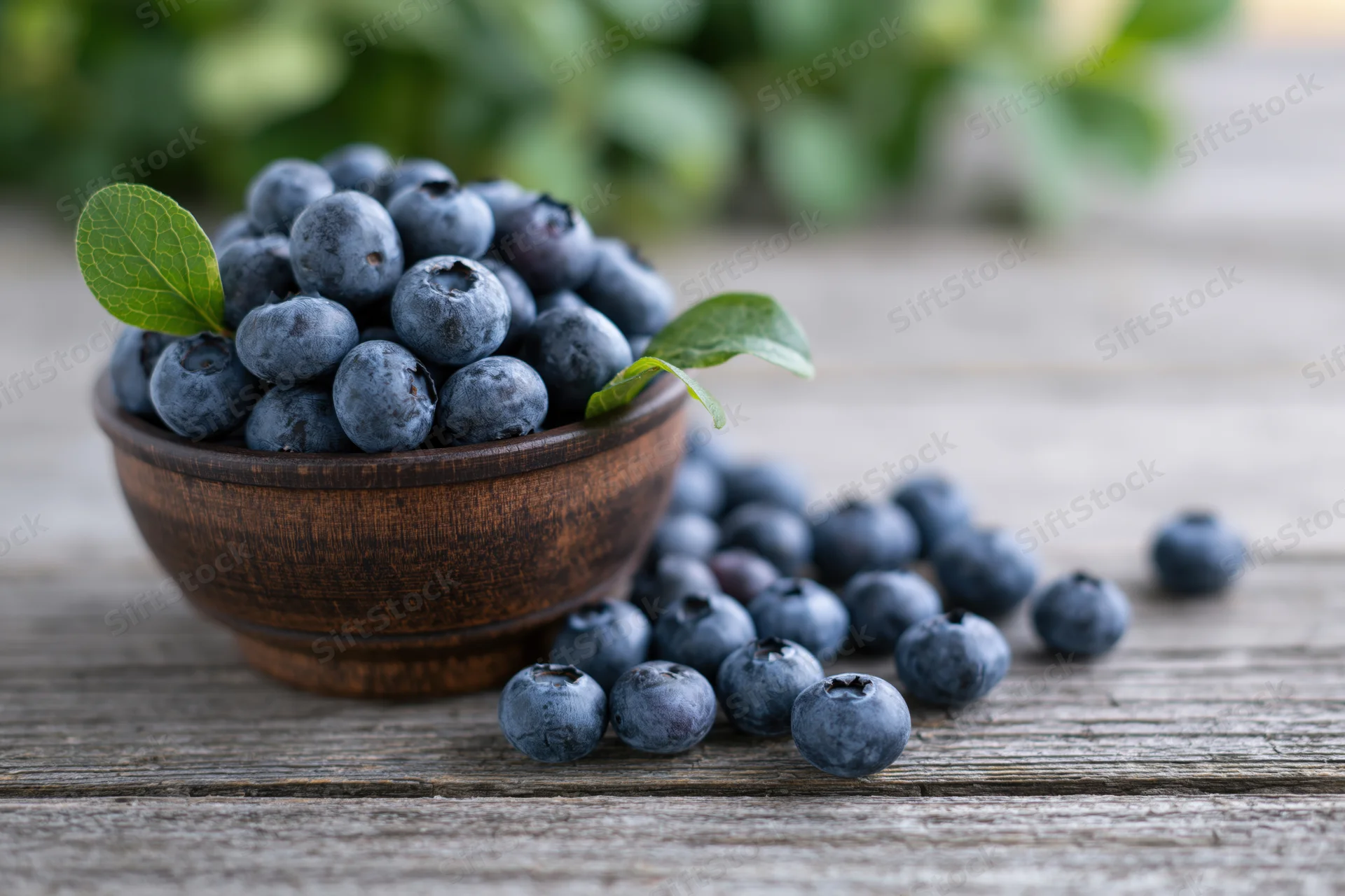 Fresh blueberries in a dark wooden bowl on a rustic wooden table with scattered berries and green leaves