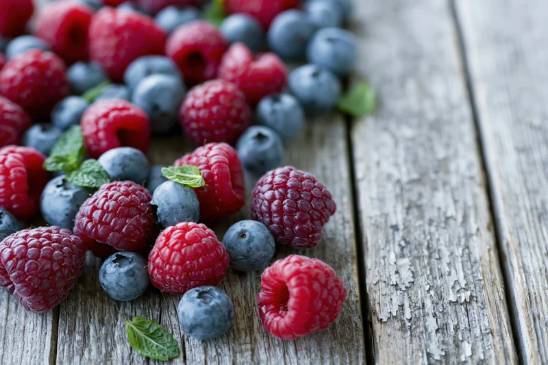 Fresh raspberries and blueberries scattered on a weathered wooden table with mint leaves