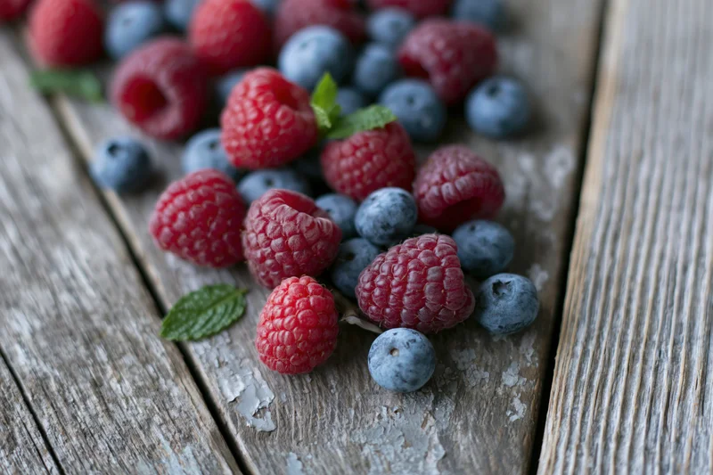 Fresh raspberries and blueberries scattered on a weathered wooden surface with mint leaves
