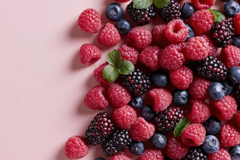 Fresh raspberries, blueberries and blackberries scattered on a pink surface with mint leaves, top-down view