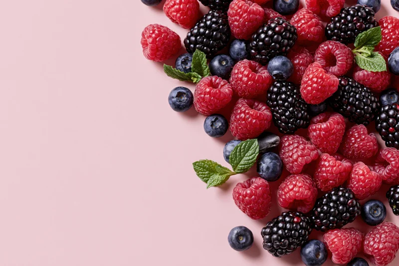 Fresh raspberries blackberries and blueberries scattered on pink surface with mint leaves and copyspace