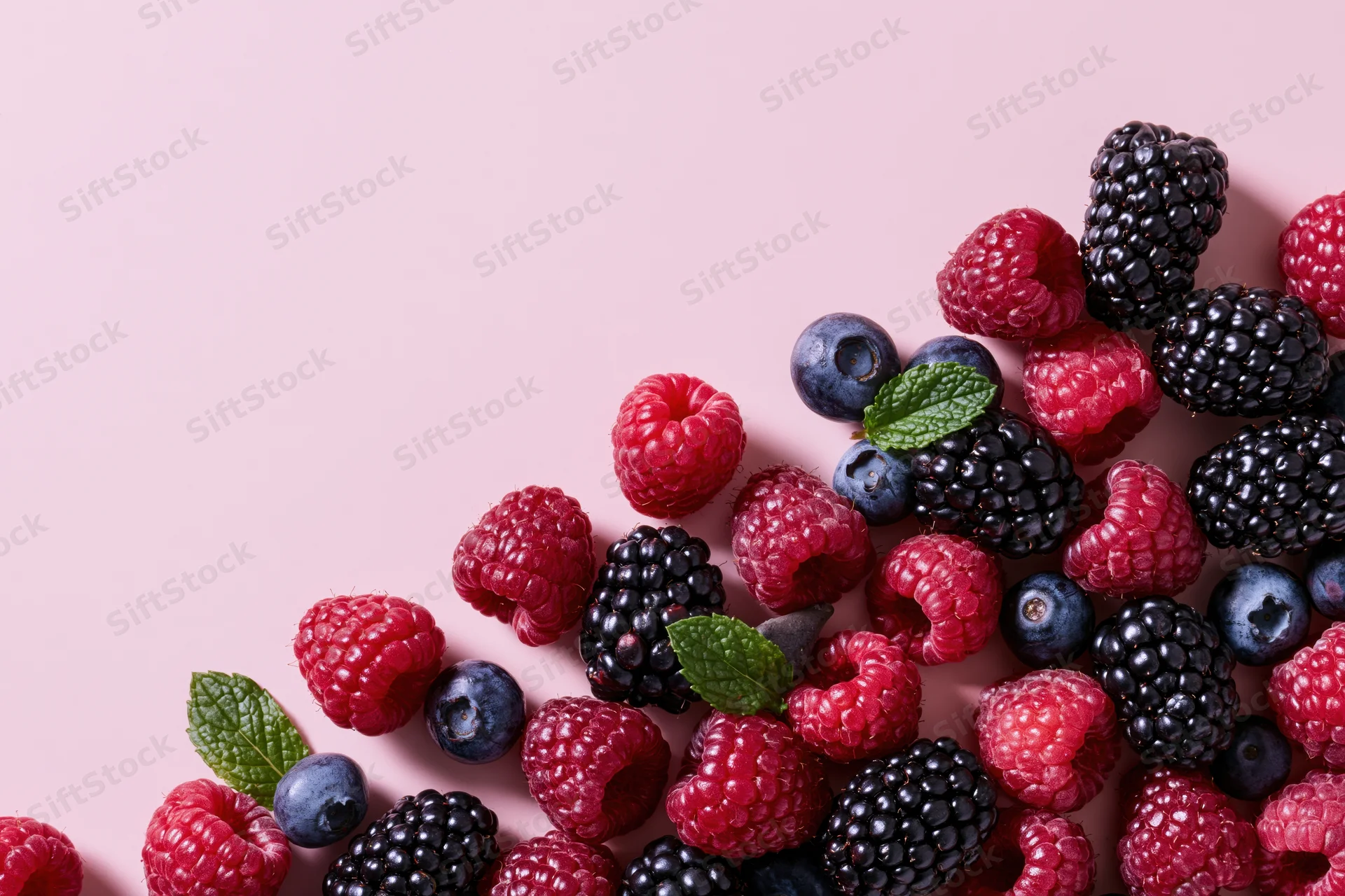 Raspberries, blackberries and blueberries scattered on a pink surface with mint leaves