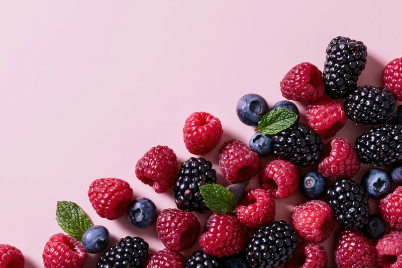 Raspberries, blackberries and blueberries scattered on a pink surface with mint leaves