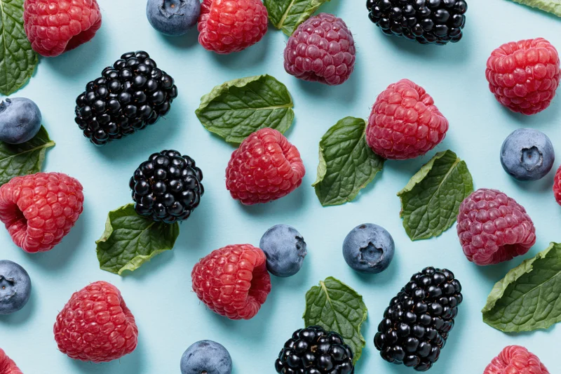 Fresh raspberries blackberries and blueberries scattered with mint leaves on a light blue surface