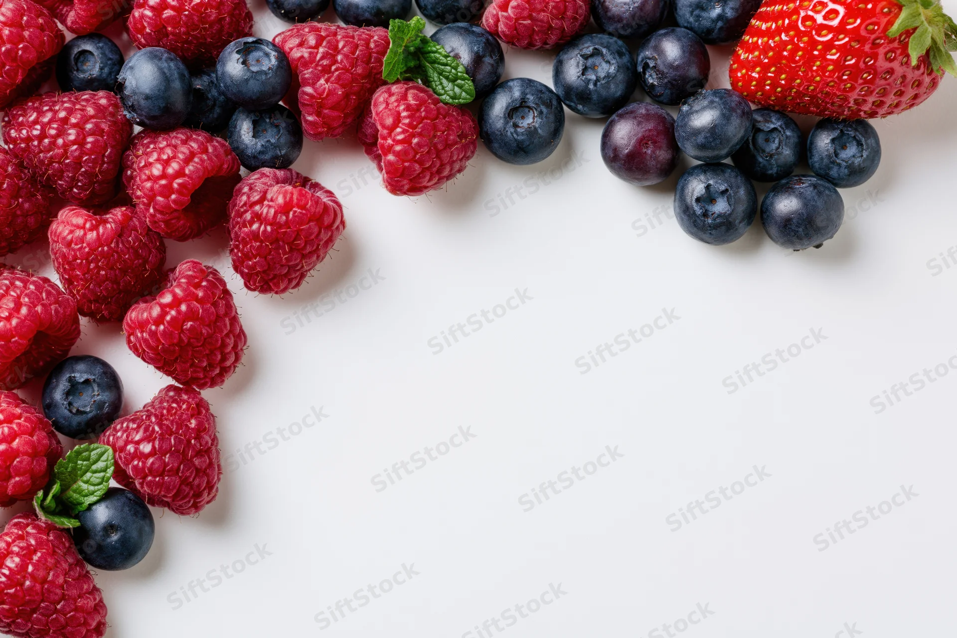 Fresh raspberries blueberries and one strawberry scattered across white surface with copyspace