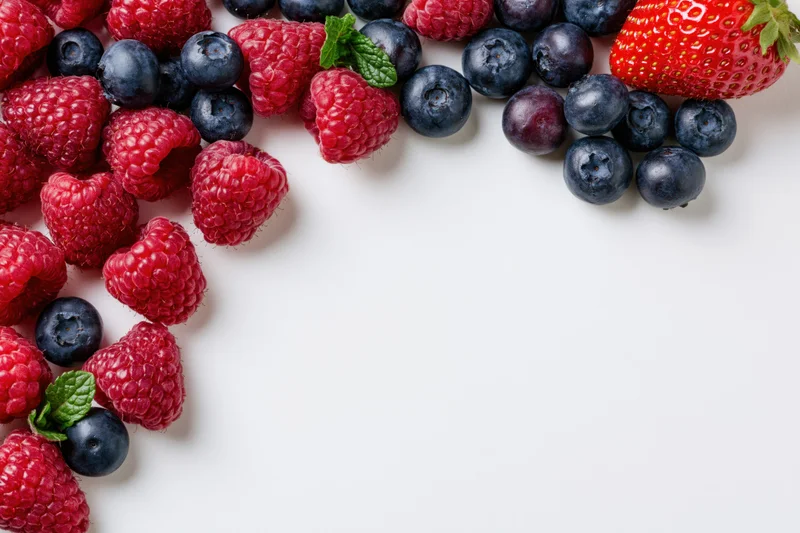 Fresh raspberries blueberries and one strawberry scattered across white surface with copyspace