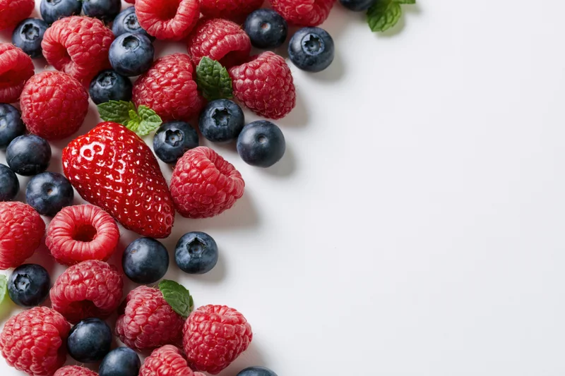 Fresh raspberries blueberries and one strawberry scattered on white surface with copyspace
