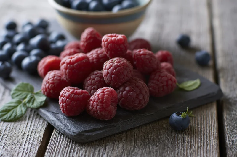 Fresh raspberries and blueberries arranged on a dark slate board with mint leaves on a wooden table