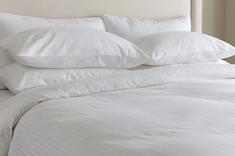 Freshly made hotel bed with three white pillows stacked against a beige headboard, covered in crisp white linens and a striped duvet