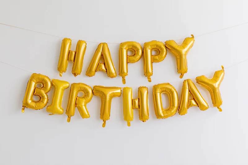 Gold foil letter balloons spelling happy birthday in two rows hanging on a string against a white wall