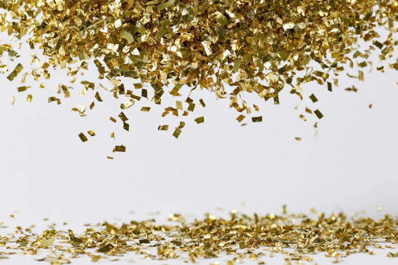 Gold metallic confetti pieces falling and scattering mid-air over white surface