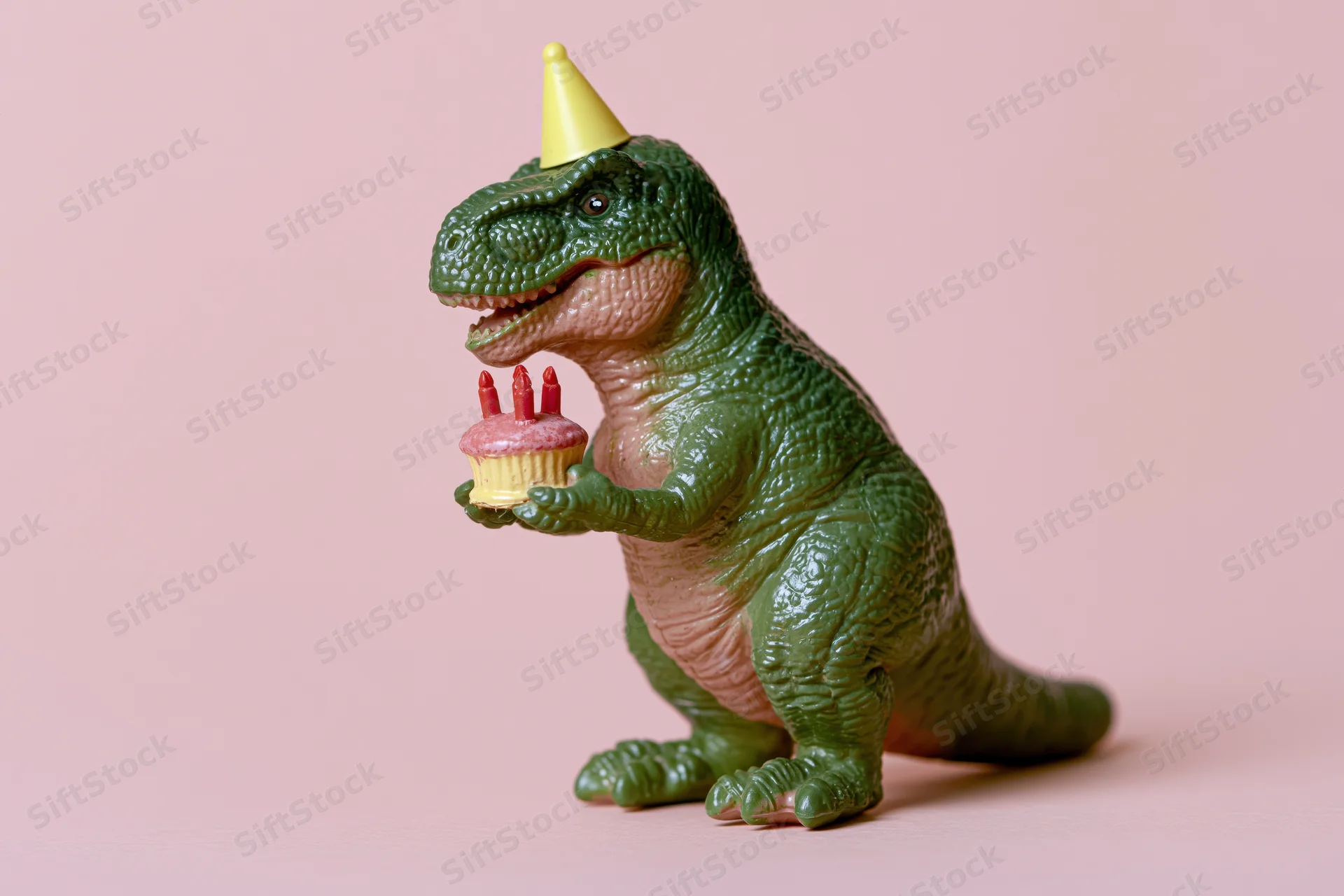 Green plastic dinosaur toy wearing yellow party hat holding cupcake with candles on pink background