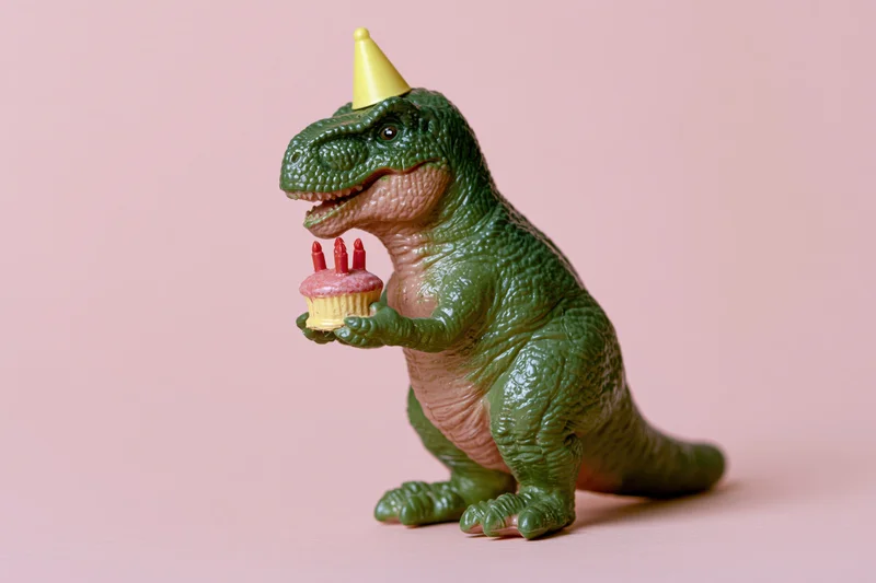 Green plastic dinosaur toy wearing yellow party hat holding cupcake with candles on pink background