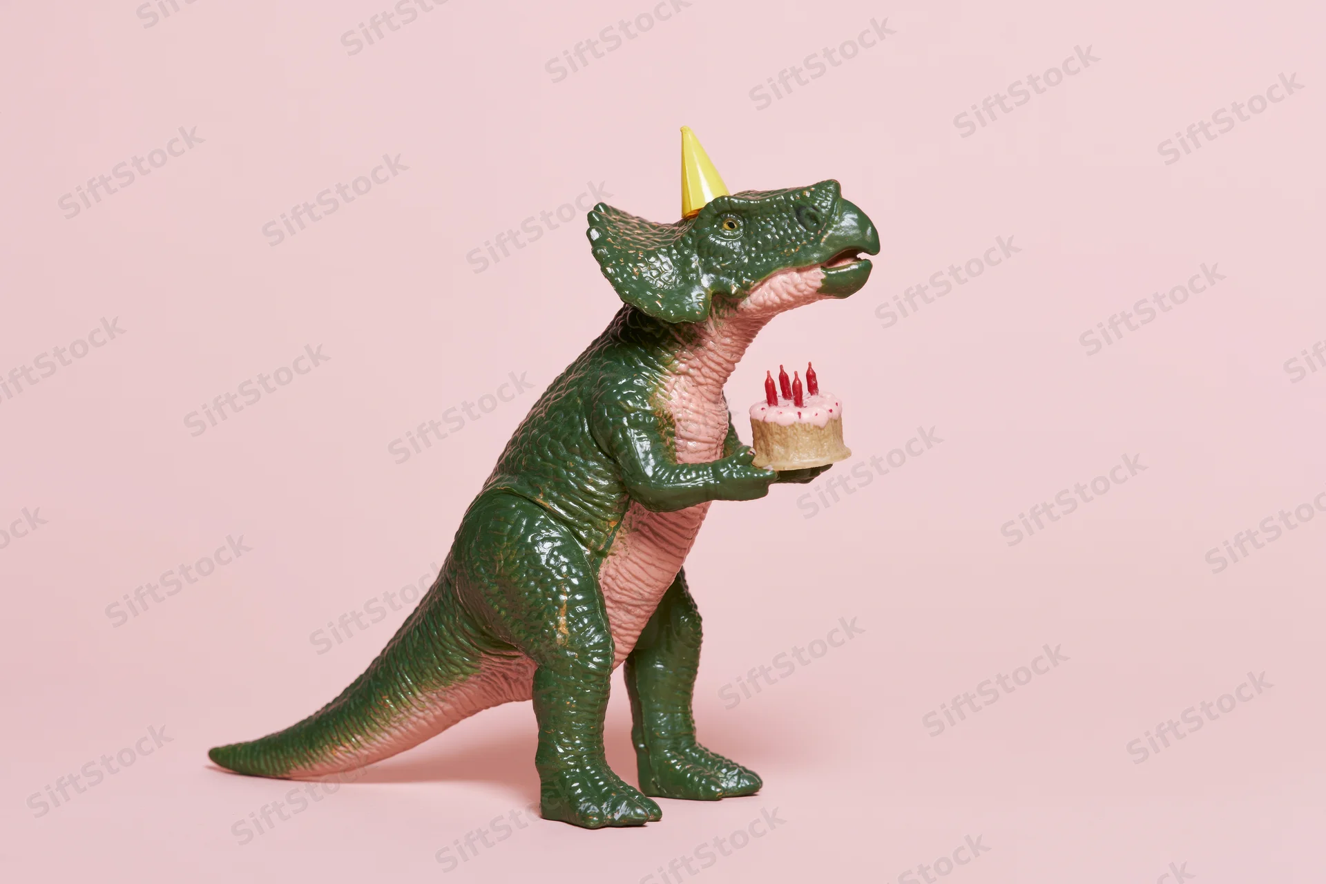Green plastic dinosaur toy wearing yellow party hat holding birthday cake with candles on pink background
