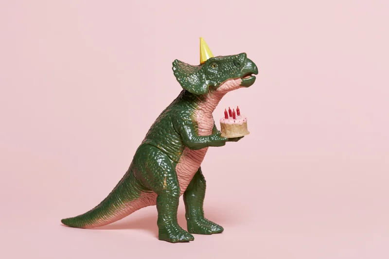 Green plastic dinosaur toy wearing yellow party hat holding birthday cake with candles on pink background