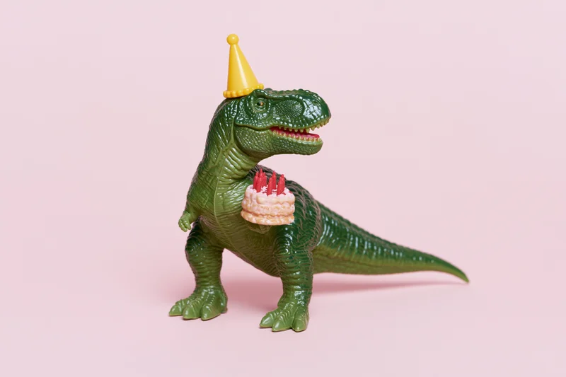 Green plastic toy dinosaur wearing yellow party hat holding birthday cake with candles on pink background