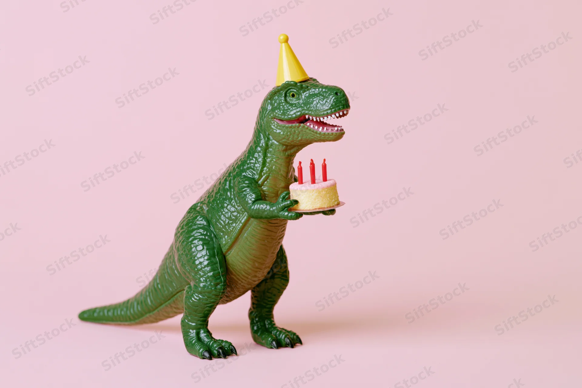 Green plastic toy dinosaur wearing yellow party hat holding birthday cake with candles on pink background