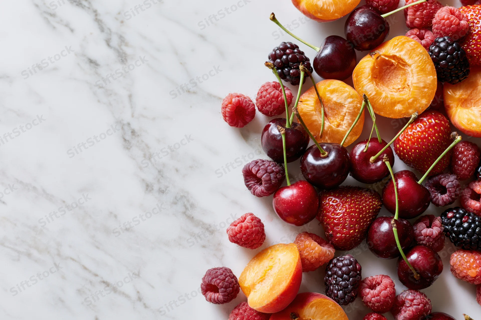 Halved apricots, cherries, strawberries, raspberries and blackberries arranged on a marble countertop with copyspace on the left